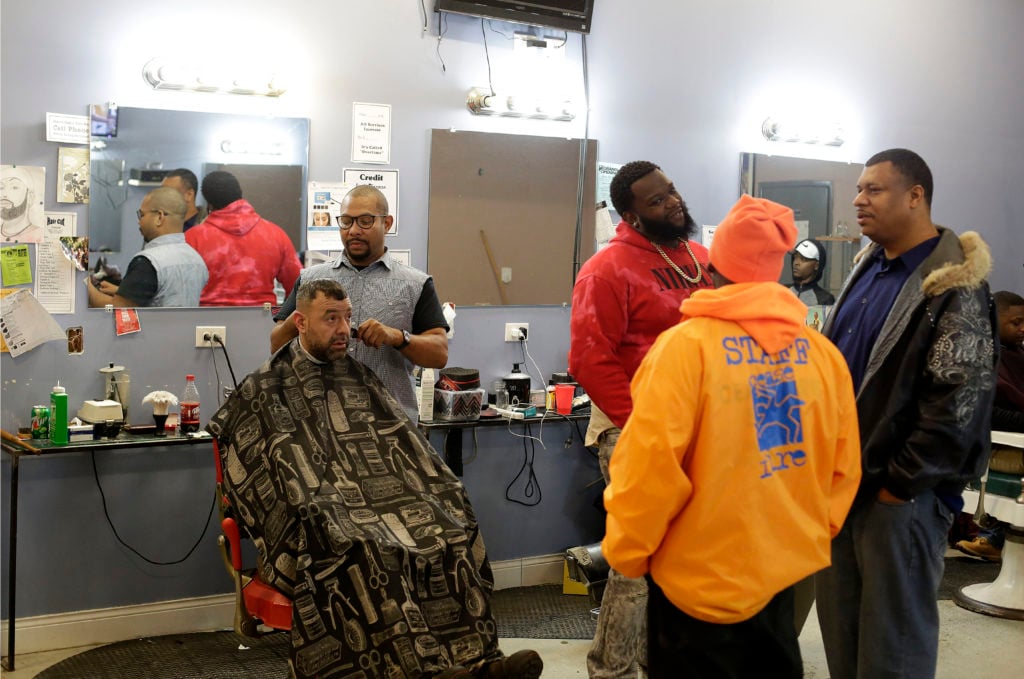 Barber Dana Dane cuts a clients hair as other customers are seen talking with Target Area supervisor Jowwaine Washington (orange jacket) January 14, 2017 in Chicago, Illinois. Ceasefire violence interrupters and Target Area workers try and prevent violent crimes such as shootings before they happen by communicating with residents in the neighborhood, gang members and relatives of gang members. (Photo by Joshua Lott for The Trace)
