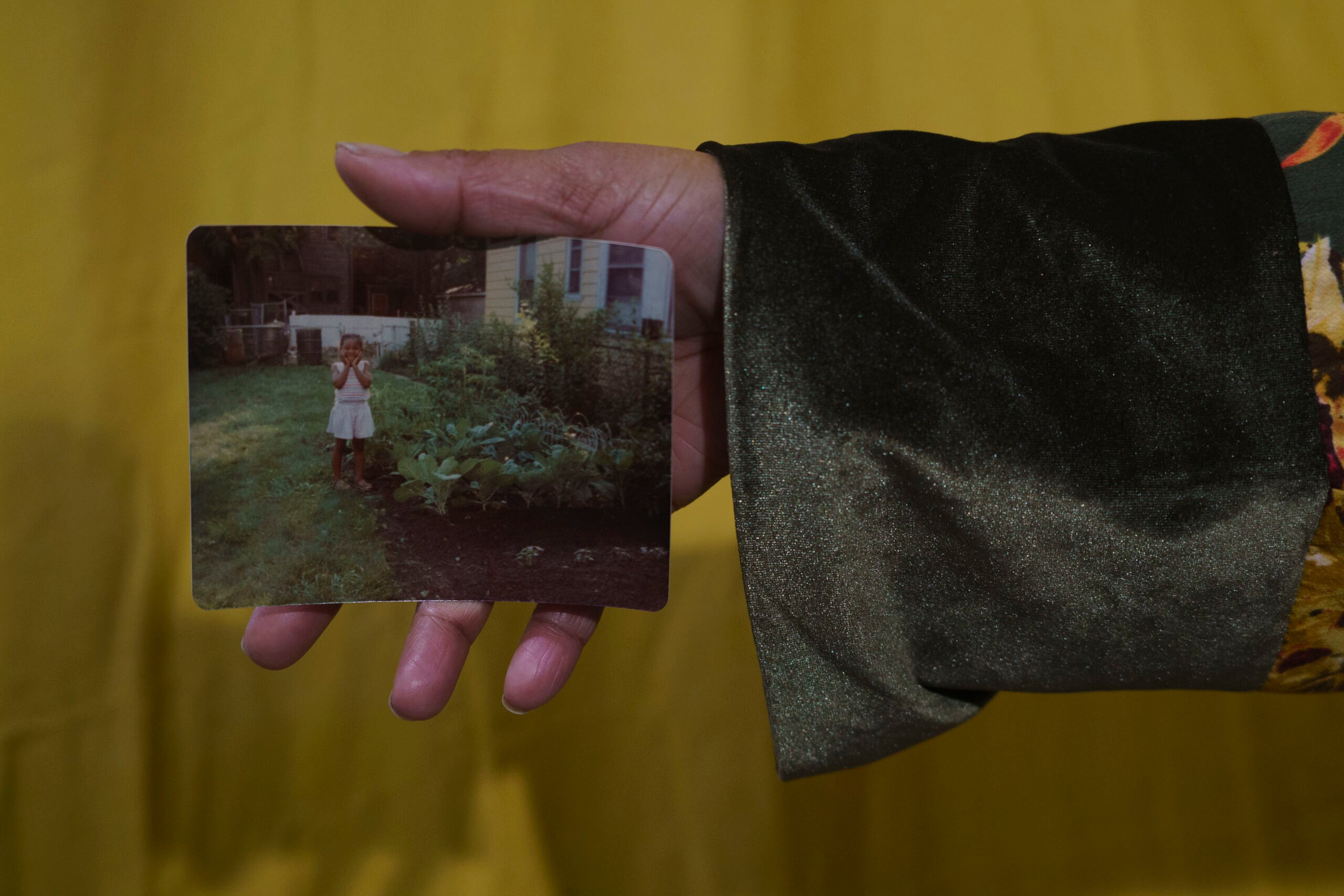 A person's hand holds a photograph print with rounded corners, showing a child standing next to a garden