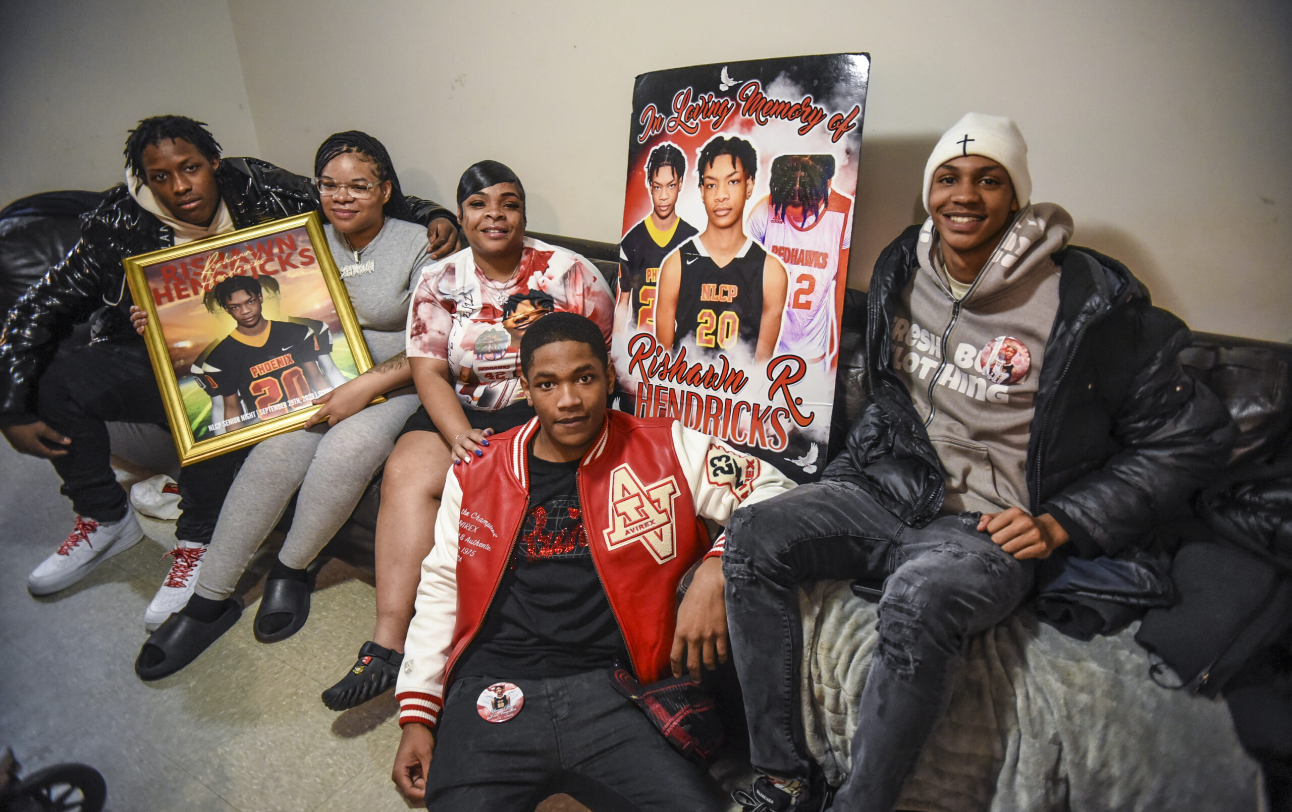 A family of five sits on a couch together, flanked by large photos of a young man. One of them reads